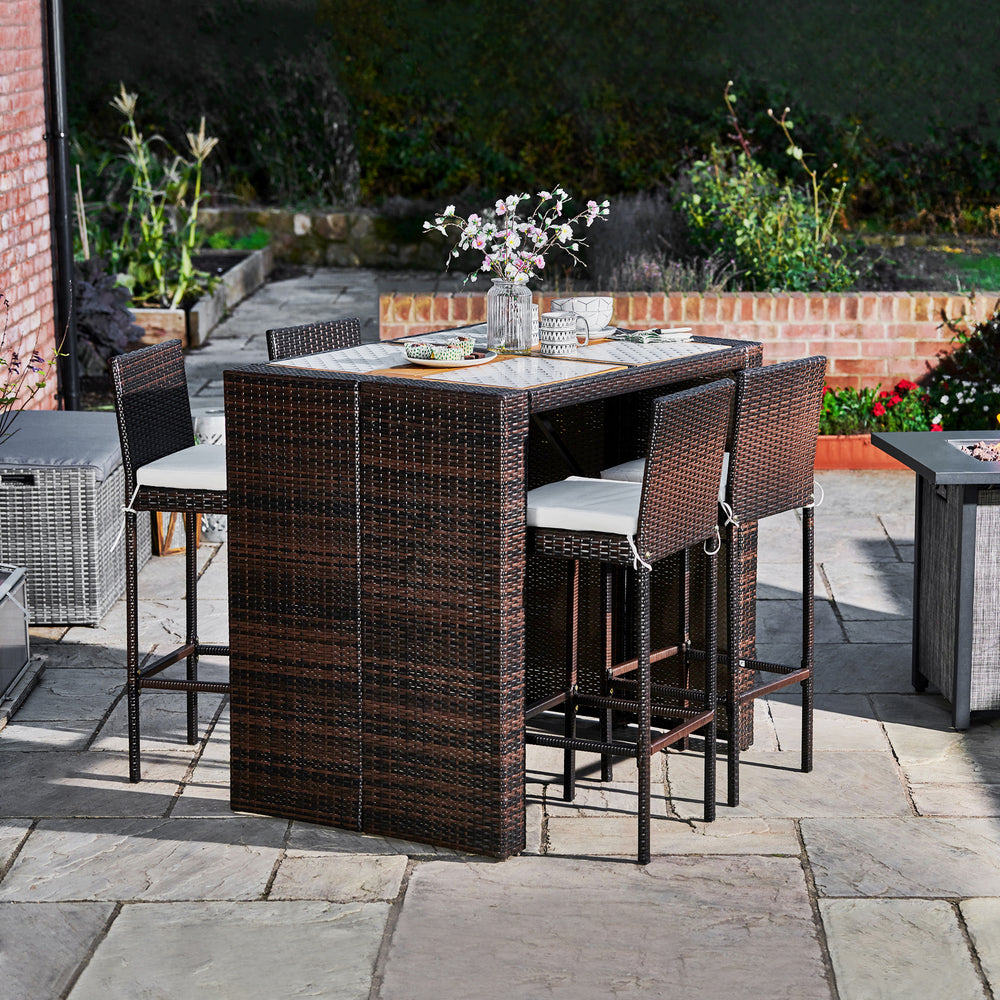 A Teamson Home Outdoor Brown PE Rattan & Acacia 5-Piece High Top Table & Stools with White Cushions on a slate patio