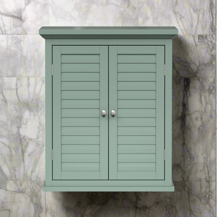 A sage green wall cabinet with silver knobs and two faux louvered doors on a marble tile wall.