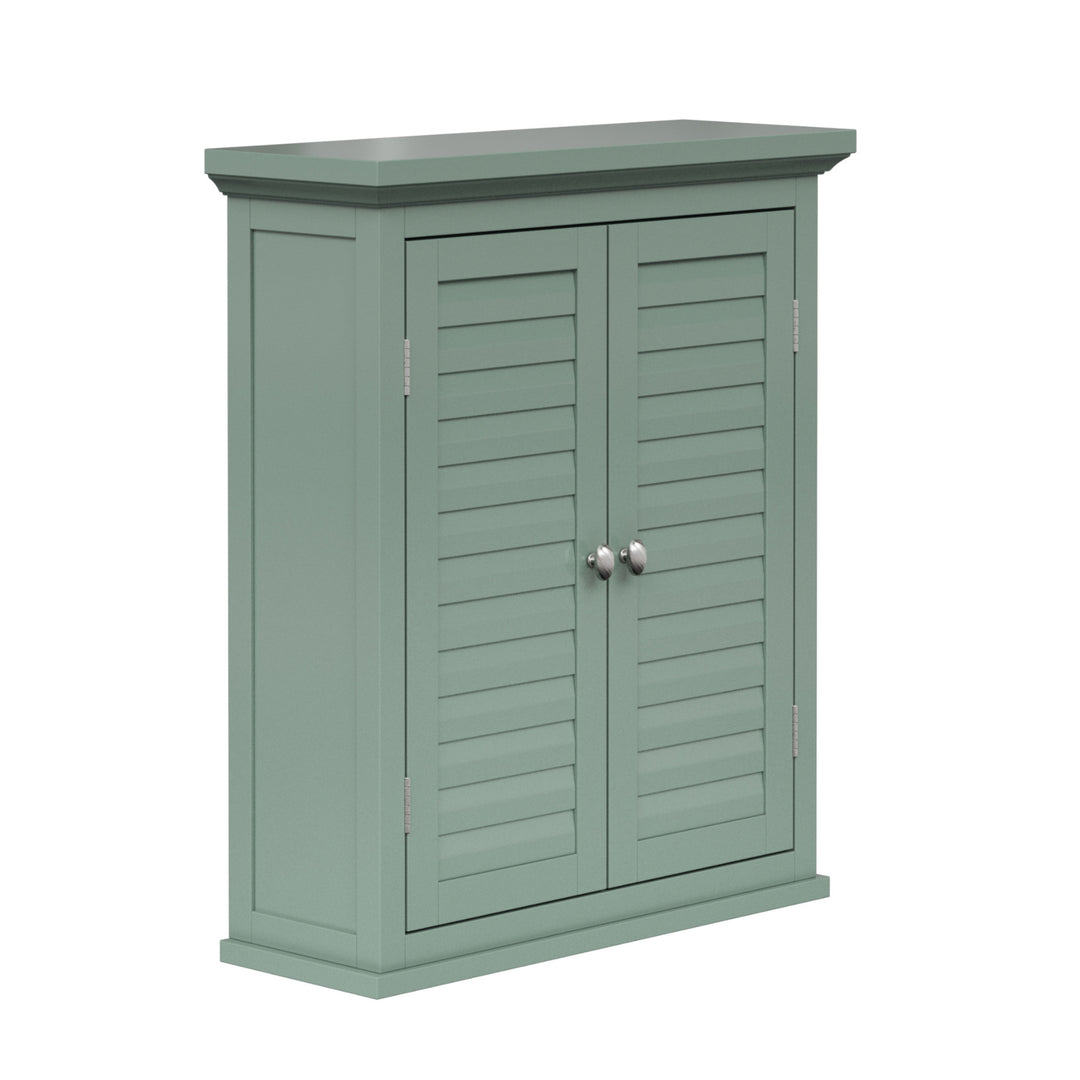 Angled view of the front of a sage green wall cabinet with chrome knobs and two faux louvered doors.