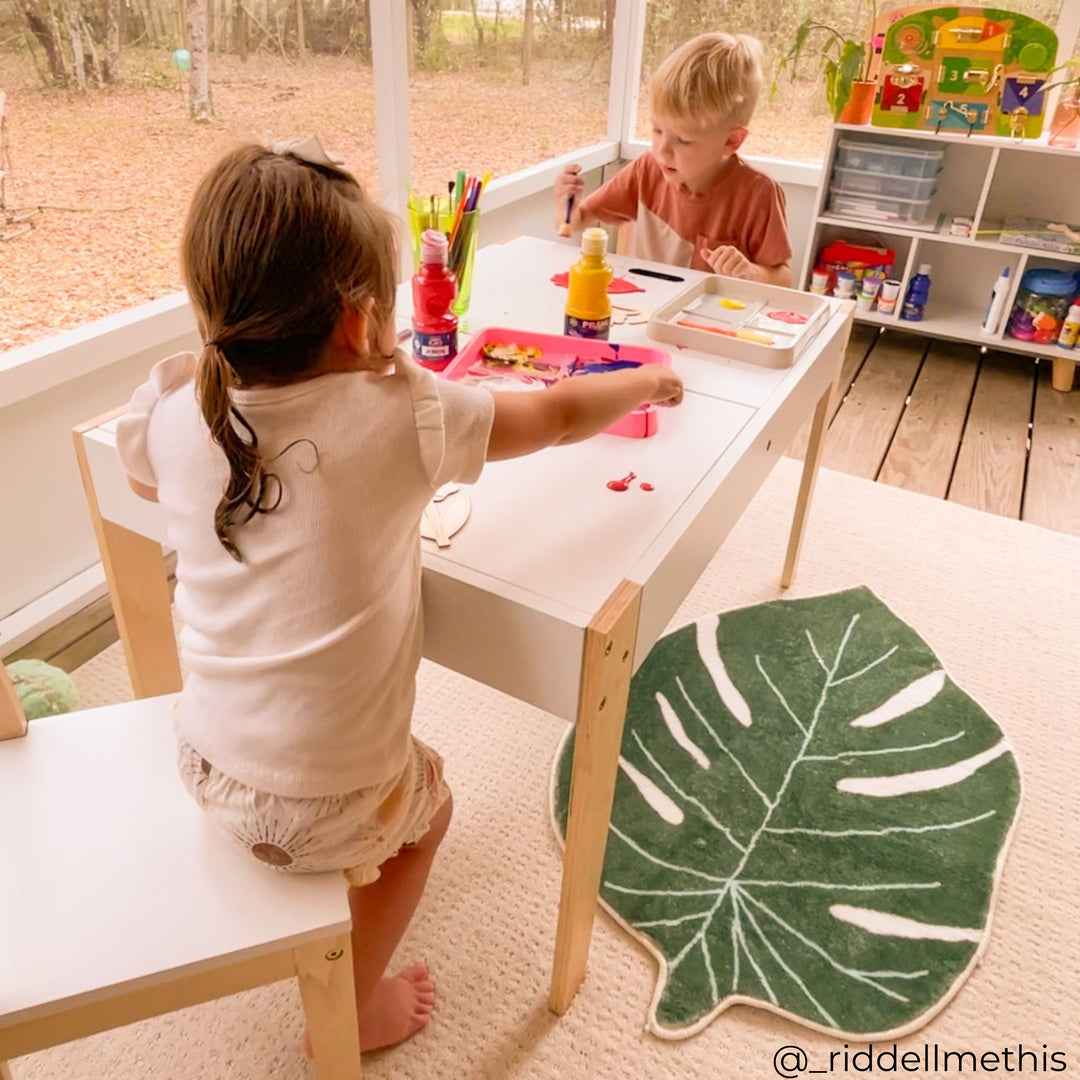 A girl and boy sit at opposite ends of a white table working on art projects.