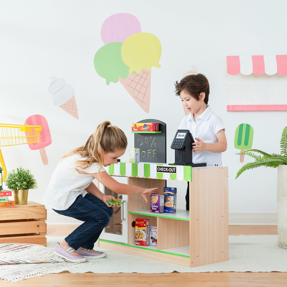 Two kids playing market stand with a Teamson Kids Cashier Austin Play Market Checkout Counter and pretend food items.