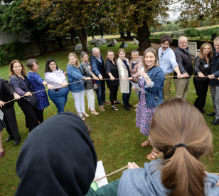 Lots of people in a circle outside on grass holding rope with a speaker in the centre.